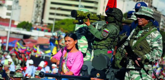 A woman in t-shirt and jeans holding an AK-47 rifle rides in a tank alongside soldiers in fatigues, one holds a rocket-launcher