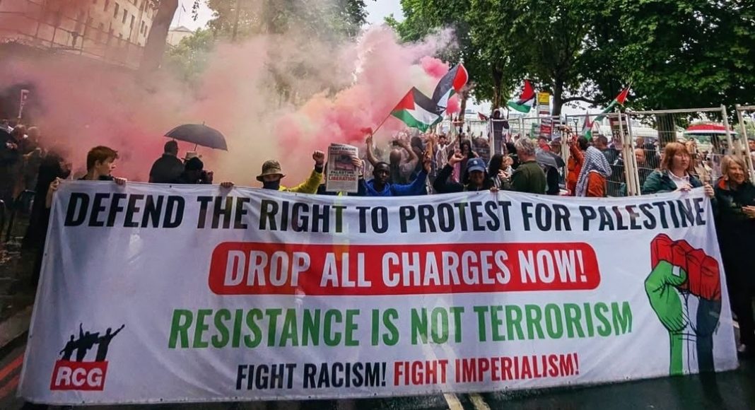 Protesters march on a London street carrying a banner reading 'defend the right to protest for Palestine/drop all charges now/resistance is not terrorism/Fight Racism! Fight Imperialism!'.