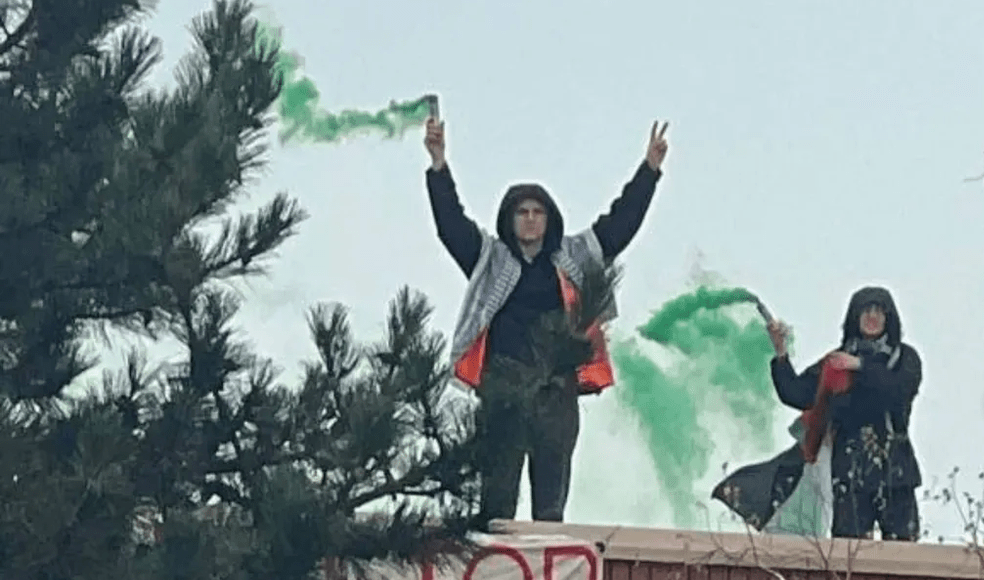 People stand with green smoke bombs on a roof with a slogan banner. Photo: People Against Genocide